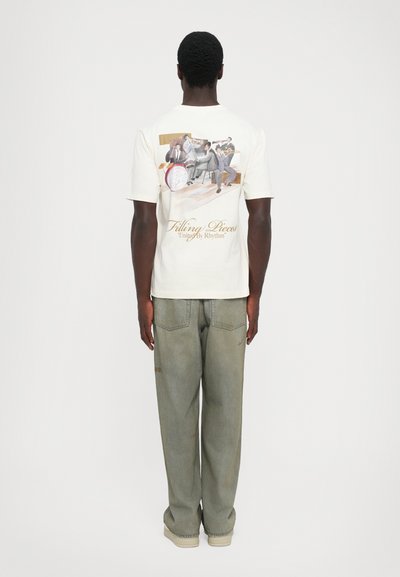 Man wearing white T-shirt with a colorful jazz band illustration and text "Filling Pieces United By Rhythm" on the back, paired with loose gray pants.
