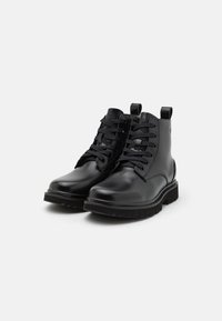Botines de cuero negro con puntera redonda, suelas planas y diseño de cordones. Presentan tiradores en el talón y suelas de goma texturizadas.