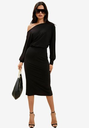 Schwarzes Off-Shoulder-Kleid mit langen Ärmeln, figurbetonter Silhouette, glattem Stoff und Knöchelriemchen-Absätzen. Das Model hält eine schwarze Handtasche.
