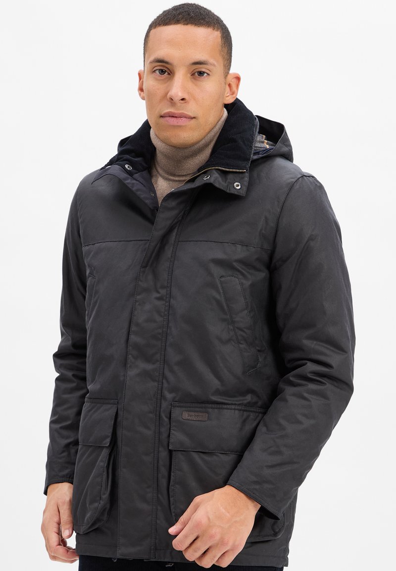 Mężczyzna w czarnej kurtce z kapturem na beżowym swetrze z golfem na jednolitym białym tle.