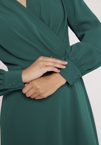 Sukienka z długim rękawem w głębokim zielonym kolorze, z zawiązanym fasonem, marszczonymi mankietami z guzikami oraz gładką, płynącą teksturą.