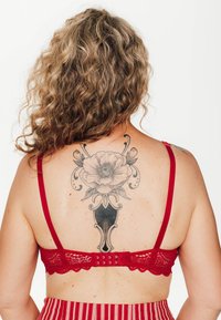 Bralette in pizzo rosso con spalline regolabili, bordi ondulati e chiusura posteriore con gancio e occhiello. Tatuaggio a forma di fiore visibile sulla schiena.