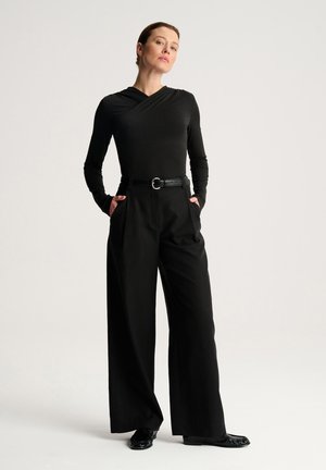 Kobieta stojąca z rękami w kieszeniach, ubrana w czarną bluzkę z długim rękawem, szerokie spodnie z paskiem oraz czarne buty na jednolitym jasnym tle.