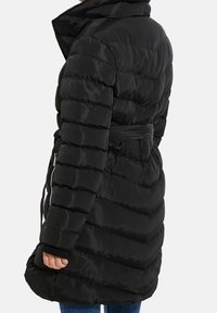 Persona vestida con una chaqueta de plumón larga y acolchada negra, con un cinturón en la cintura y un bolsillo lateral con cremallera, jeans azules visibles debajo de la chaqueta.