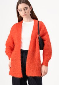 Lys oransje, fluffy, langermet cardigan laget av mykt materiale, åpen frontdesign, brukt over en hvit topp, kombinert med svarte bukser.