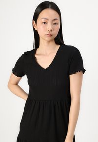 Black dress crafted from ribbed fabric, featuring a V-neck and short ruffled sleeves, with a gathered waistline and flowing bottom.