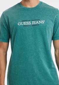 Teal bombažna majica s kratkimi rokavi in krožnim izrezom. Spredaj je prikazan napis "GUESS JEANS" v belem in črnem. Teksturen zaključek.