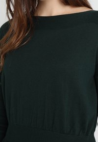 Mörkgrön stickad tröja med bred bateau-halsringning, ribbad nederkant och mjuk textur. Har långa ärmar och en sammanknuten midjedetalj.
