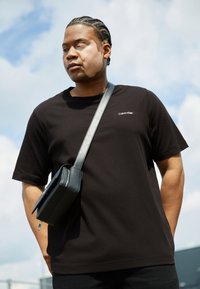Sort bomulds T-shirt med korte ærmer, der har et lille hvidt Calvin Klein-logo. Kombineret med en struktureret sort crossbody taske.