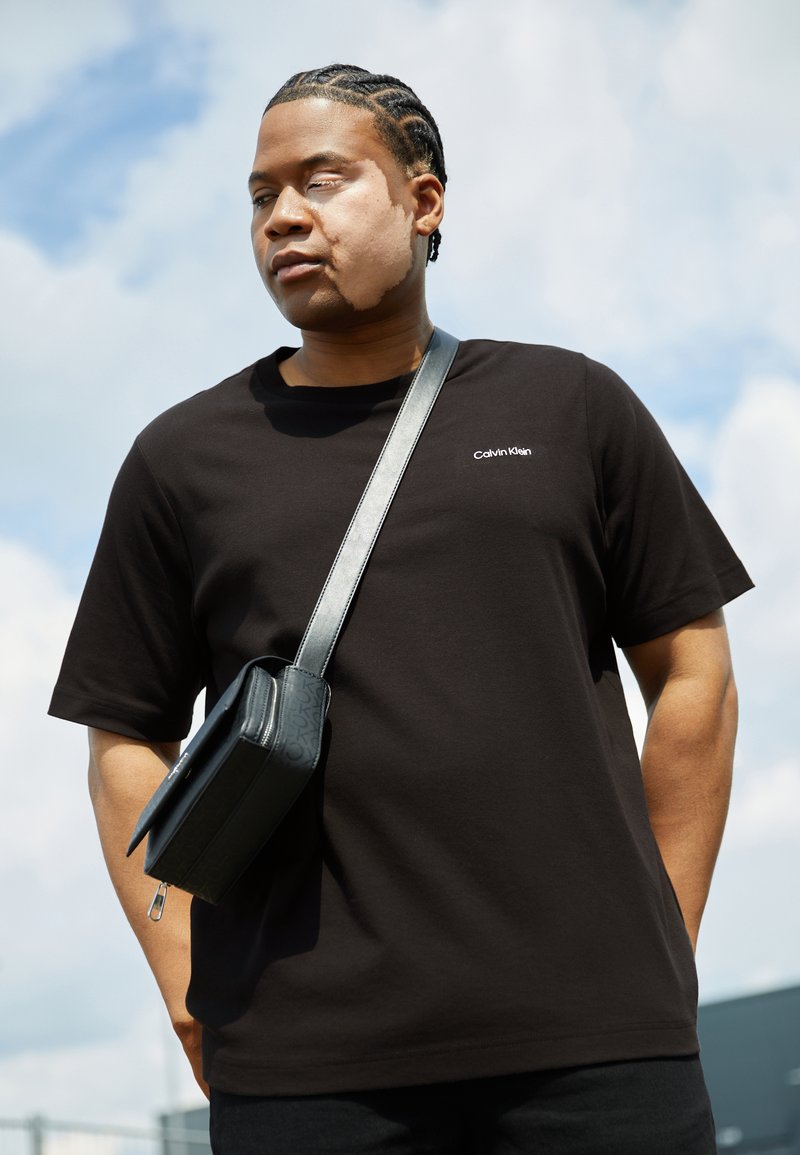 Sort bomulds T-shirt med korte ærmer, der har et lille hvidt Calvin Klein-logo. Kombineret med en struktureret sort crossbody taske.
