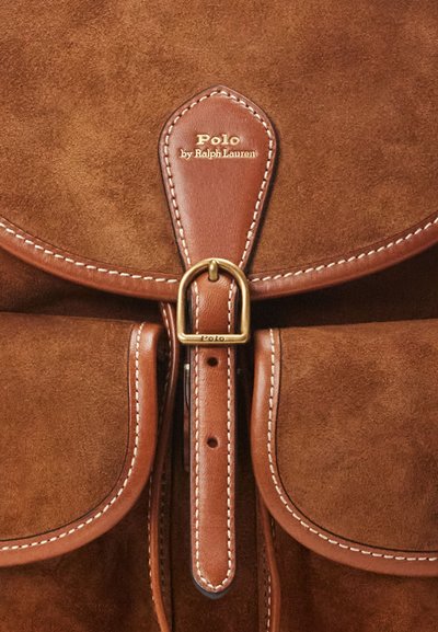 Hnedý semišový batoh s koženými akcentmi, obsahujúci zlatú pracku a biele šitie. Logo "Polo by Ralph Lauren" je výrazne zobrazené.