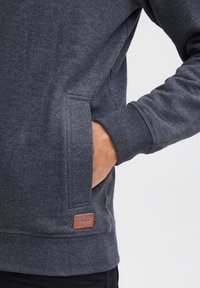 Sudadera de forro polar gris con bolsillo tipo canguro y puños acanalados. Etiqueta marrón claro con la inscripción "BLEND" cosida en el dobladillo inferior.