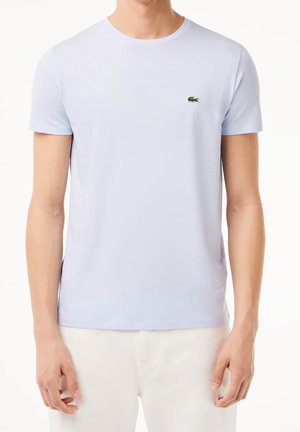 Homme portant un t-shirt bleu clair à manches courtes avec un petit logo crocodile vert sur la poitrine gauche et un pantalon écru.