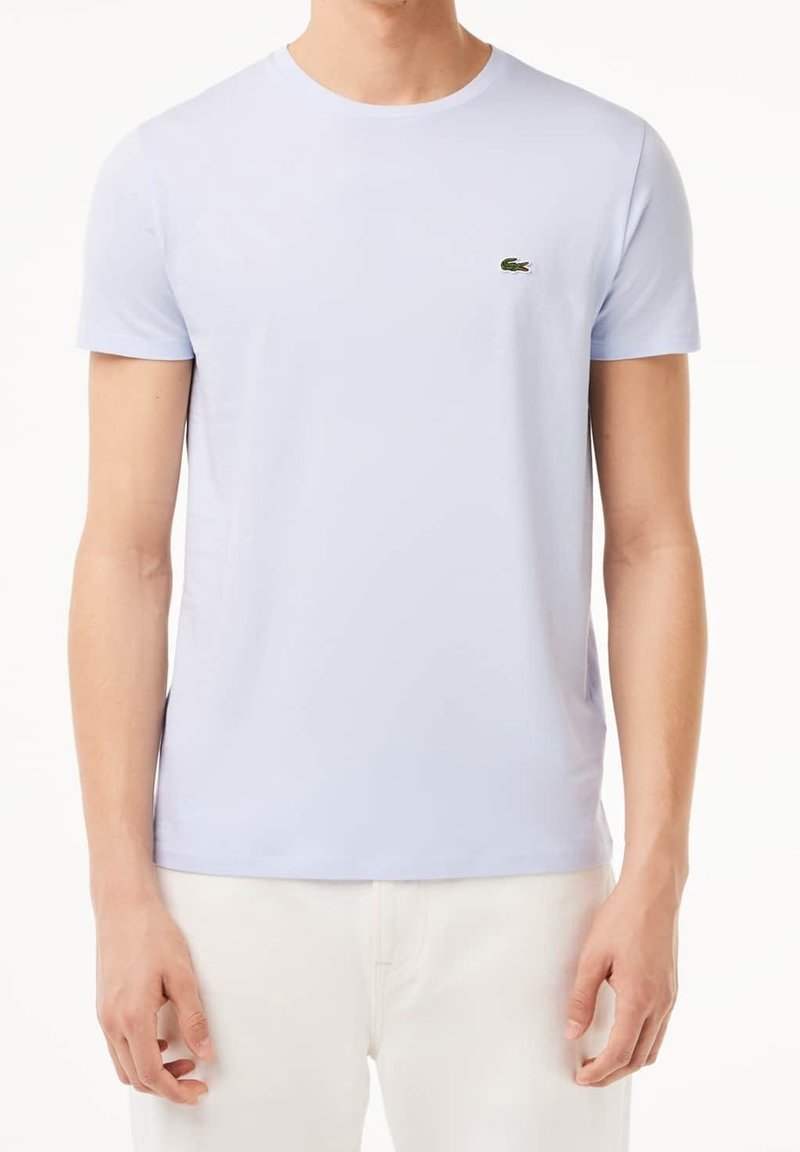 Homme portant un t-shirt bleu clair à manches courtes avec un petit logo crocodile vert sur la poitrine gauche et un pantalon écru.