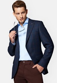 Granatowa marynarka w kratę z brązowymi akcentami, jasnoniebieska koszula i brązowe spodnie. Marynarka ma klapy w kształcie notcha i dwa guziki. Okulary trzymane w ręku.