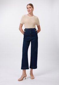 Beige gebreide top met textuur, gecombineerd met hooggekraagde donkere denim wijde broek met gouden accenten en een korte manchette. Minimalistische bandjeshakken.