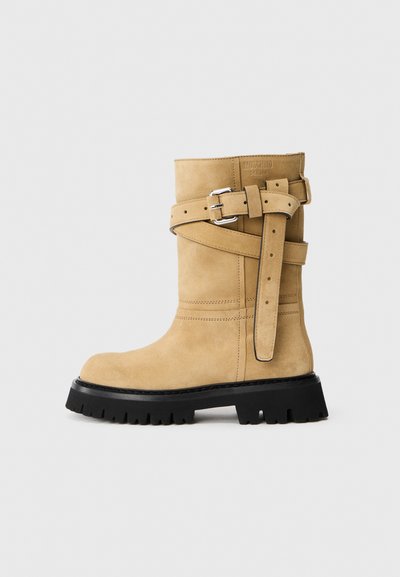 Tan suède laarzen tot de kuit met een zwarte chunky zool, zilveren gespbanden die kruisen aan de zijkant, en een gegraveerd Moschino Milano logo nabij de bovenkant.