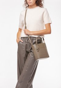 Piccola borsa a tracolla verde oliva con un design strutturato, superfici lisce e texture, e una tracolla removibile, tenuta da una persona.