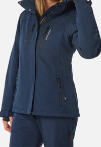 Femme portant une veste et un pantalon bleu marine avec plusieurs poches zippées, ajustant sa capuche ou son col de la main droite.