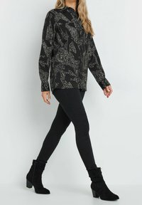 Schwarzes Langarmshirt mit abstraktem weißen Muster, Knopfverschluss und lässiger Passform, kombiniert mit schwarzen Leggings und lockeren Knöchelstiefeln.