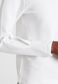 Bílá dlouhá rukávová košile s ribovanou texturou, s rovný lem a jemnou, hladkou látkou. Oblast zápěstí je zobrazená s přiléhavým manžetou.