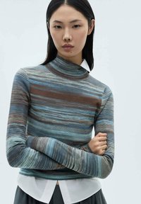 Långärmad topp med hög krage, prydd med horisontella ränder i blått, brunt och beige. Bärs över en vit skjorta med krage.