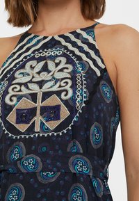 Halterkleid mit einem dunkelblauen gemusterten Stoff, kunstvollen Blumen- und geometrischen Stickereien in Creme und Lila sowie einer gebundenen Taille.