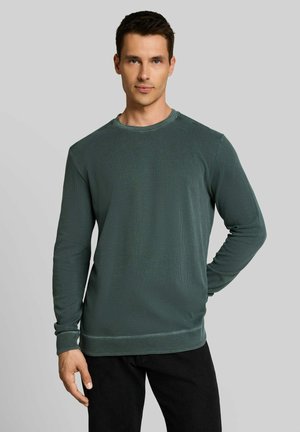 Mann trägt ein dunkelgrünes, strukturiertes Hemd mit langen Ärmeln und schwarze Hose, steht mit einer Hand hinter dem Rücken vor einem einfarbigen Hintergrund.