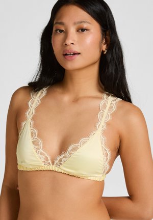 Junge Frau mit langen dunklen Haaren, die ein hellgelbes Bralette mit Spitzenbesatz trägt und vor einem schlichten hellen Hintergrund posiert.