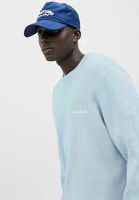 Light blue sweater with a textured knit, featuring a round neckline and subtle embroidered branding. Navy blue cap with white text accents.