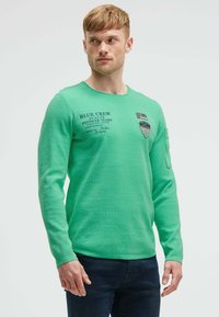 Grünes Langarmshirt mit einem runden Ausschnitt, versehen mit bedrucktem Text und Patches auf der Brust und dem Ärmel. Kombiniert mit dunkelblauen Jeans.