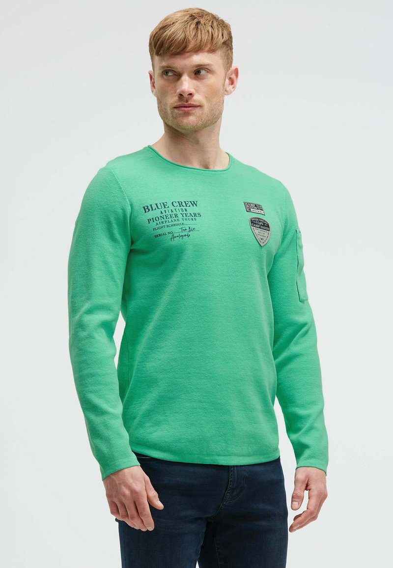 Grünes Langarmshirt mit einem runden Ausschnitt, versehen mit bedrucktem Text und Patches auf der Brust und dem Ärmel. Kombiniert mit dunkelblauen Jeans.