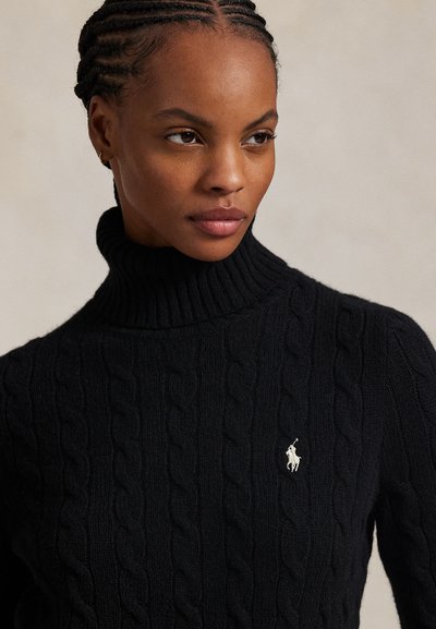 Giovane donna con i capelli intrecciati che indossa un maglione nero a collo alto lavorato a treccia con un piccolo logo bianco sul petto.