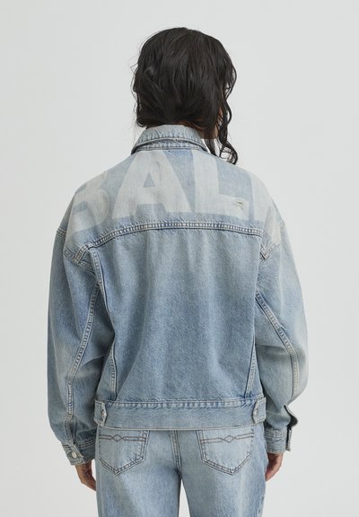 Person with dark hair wears light blue denim jacket and jeans, facing away, showing faded large letters on jacket back.