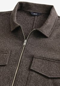 Ruda tekstūruota zip-up striukė su apykakle, turinti dvi krūtinės kišenes ir sidabrinį metalinį užtrauktuką. Prekės ženklo "NEXT" etiketė matoma viduje.