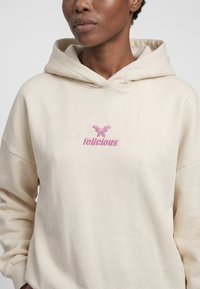 Persoon draagt een lichtbeige hoodie met een roze vlinder en het woord "felicious" geborduurd op de borst.