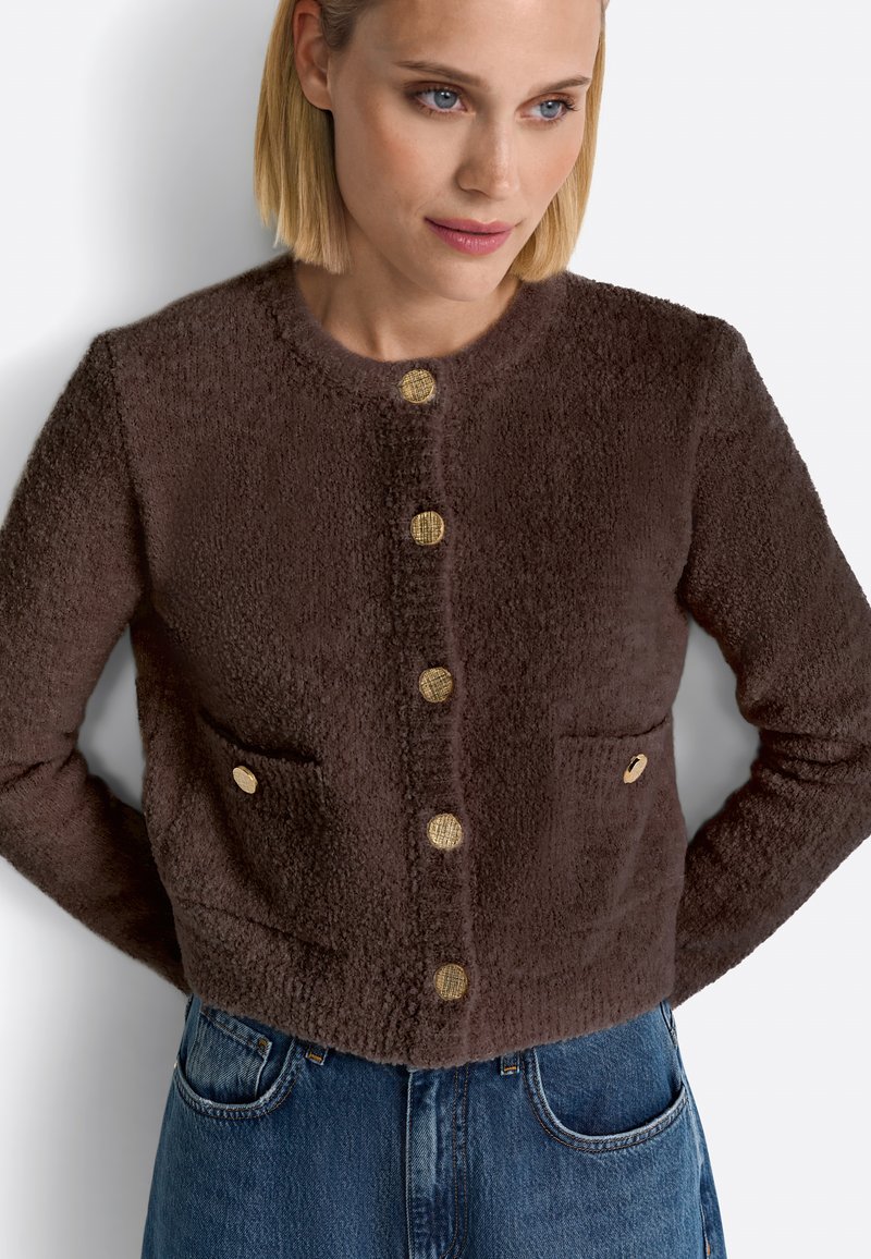 Giacca marrone con texture, colletto rotondo, bottoni frontali dorati e due tasche anteriori. Indossata con jeans di denim.