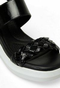 Sandalia negra con una correa ancha y brillante y una banda frontal trenzada. Presenta una plantilla negra texturizada y una suela de plataforma blanca.