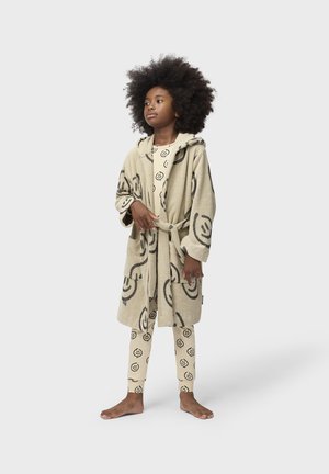 Barn med stort krøllet hår iført beige pyjamas med smilende ansigt og en matchende hætteklæde med lommer og bælte, stående barfodet på en hvid baggrund.