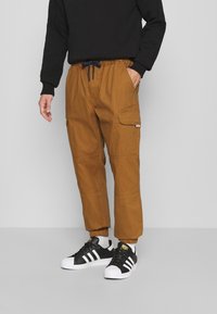 Pantalons cargo en toile marron avec une taille élastique et un cordon de serrage, dotés de poches latérales et de poignets fuselés, associés à des baskets noires.