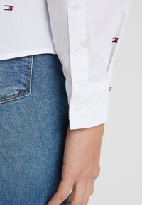 Camisa blanca de botones con mangas largas, que presenta un sutil detalle del logo cerca del puño, combinada con unos jeans de mezclilla azules.