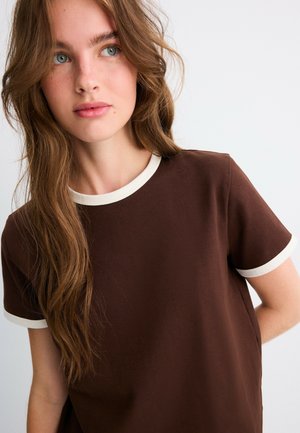 Jeune femme aux longs cheveux bruns ondulés portant un t-shirt marron foncé avec un liseré blanc au niveau de l'encolure et des manches, regardant légèrement sur le côté.