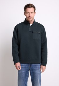 Uomo che indossa un pullover scuro con un quarto di bottone e una tasca sul petto, accompagnato da jeans blu, in piedi davanti a uno sfondo chiaro e semplice.