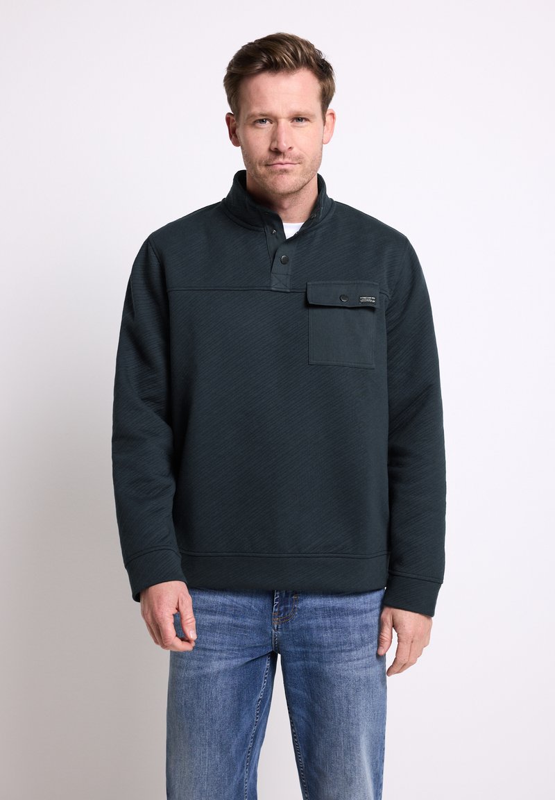 Uomo che indossa un pullover scuro con un quarto di bottone e una tasca sul petto, accompagnato da jeans blu, in piedi davanti a uno sfondo chiaro e semplice.