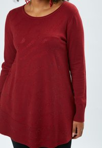 Roter Pullover mit langen Ärmeln und strukturiertem Muster, mit weitem Rundhalsausschnitt und gerippten Bündchen. Das Material wirkt weich und hat subtile Glitzereffekte.