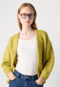 Limettivihreä neulecardigan, jossa on väljä istuvuus, avonainen etuosa ja teksturoitu kangas. Se on kerroksittain valkoisen tiukan paidan ja sinisten farkkujen päällä.