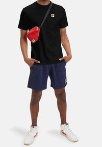 Black t-shirt with small logo, navy shorts, red sling bag, and white sneakers with red accents, featuring casual, comfortable fabric and fit.