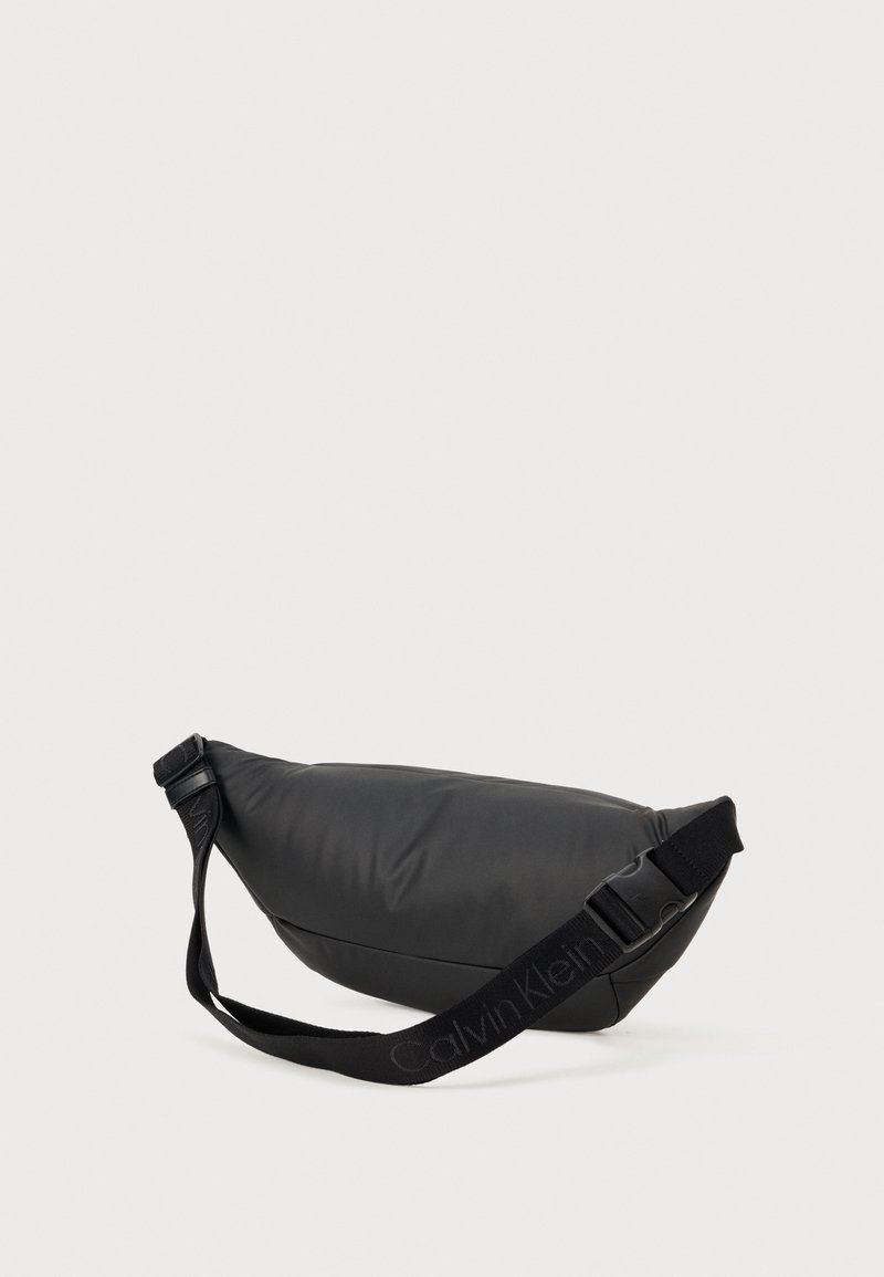 Sac bandoulière noir, à texture douce, de forme incurvée, comportant une sangle réglable avec des détails de logo et une fermeture à boucle.