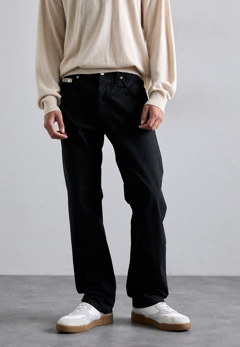 Jeans de mezclilla negros con un corte recto, llevados con un suéter de punto beige claro. Zapatillas blancas con suelas de goma complementan el atuendo.