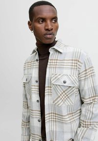 Camisa de franela a cuadros en gris claro y beige, con dos bolsillos en el pecho, un cuello y cierre de botones, llevada sobre un cuello de tortuga marrón oscuro.